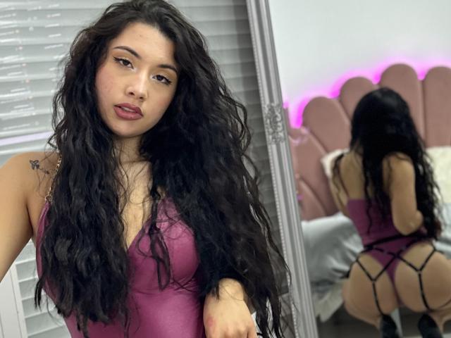 ScarlletRays with long, wavy black hair, wearing a purple lace-up bodysuit, showing her tits and ass in a mirror reflection, pink headboard, and purple lighting.