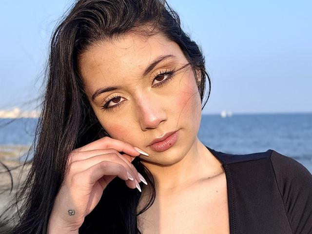ScarlletRays has long black hair, brown eyes, and full lips with a slight pout. She's wearing a black top, showing her tits and a small heart tattoo on her hand.