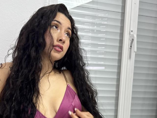 ScarlletRays with long curly black hair, wearing a purple bra, looking slightly to the side, showing her small boobs and pink lips, standing near white blinds.