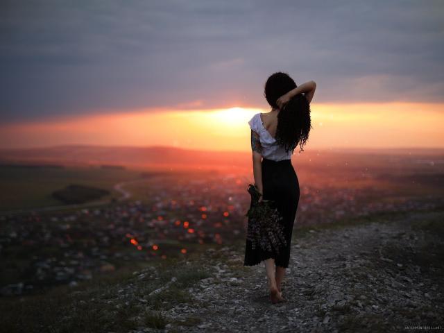 BettyStone69 stands on a rocky hill, back to the camera, in a white off-shoulder top and black skirt, with long curly hair, sunset glowing behind her.