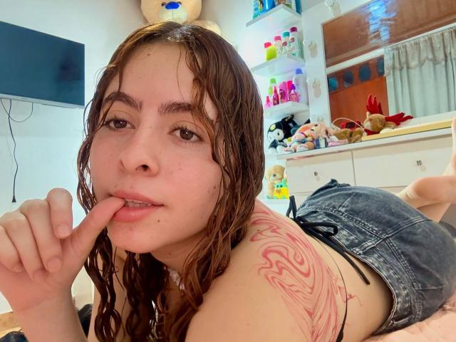 EmmaFosterX with wet brown hair, biting her finger, has a red tattoo on her right shoulder, wearing denim shorts, lying on a bed, looking at the camera.
