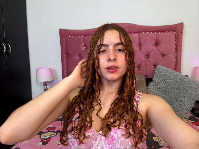 EmmaFosterX with long, curly brown hair, pink lace top, and pink bedroom, holding her head with a relaxed, slightly open-mouthed expression. Boobs are visible, medium-sized.