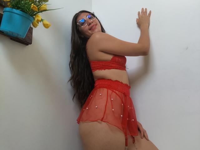 DaMikaela with long dark hair, glasses, red lace bra, and sheer red skirt with pearl details, standing against a white wall, looking back, showing her ass and tits.