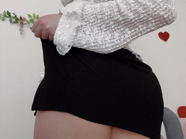 O traseiro de Rubbi está exposto sob uma saia preta, enquanto ela levanta uma blusa branca, texturizada e de mangas compridas, revelando a pele clara e um vislumbre de sua vulva..