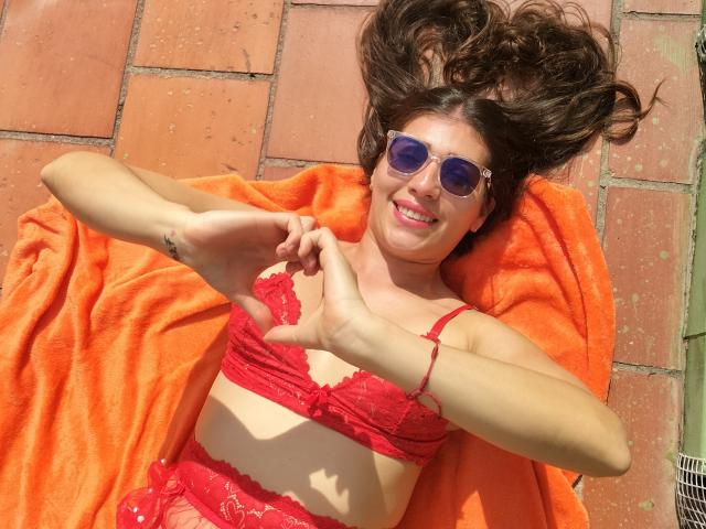 DaMikaela lying on an orange towel, wearing red lace bra and panties, with her hands in a heart shape, smiling, and wearing blue sunglasses, with her brown hair spread.