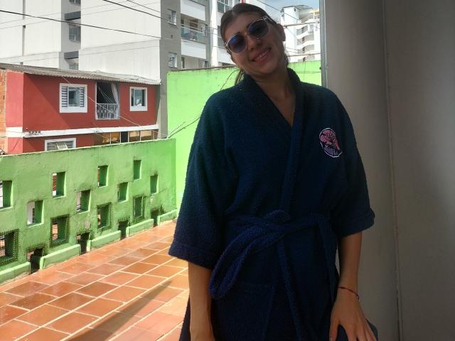 DaMikaela with dark blue bathrobe, tied at the waist, wearing sunglasses, standing on a tiled balcony, green wall behind, and red building in the background. Her ass is visible.