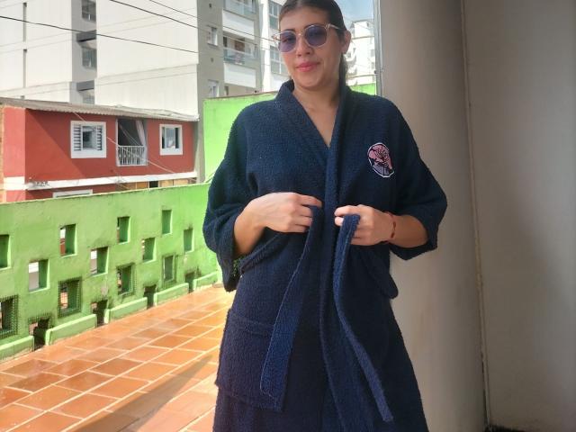 DaMikaela, wearing a dark blue, fuzzy, bathrobe with a red and white logo, standing on a tiled balcony, holding the robe's tie, with sunglasses on and a slight smile.