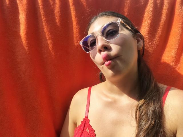 DaMikaela with pouty lips, wearing red lace top and clear sunglasses, against a bright orange wall, has long brown hair, light skin, and a cute, sexy face.