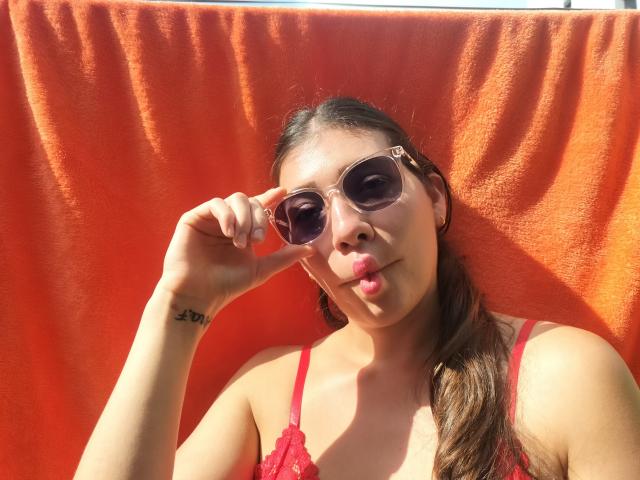 DaMikaela with brown hair in a ponytail, wearing red lace lingerie, and pink sunglasses, slightly puckering her lips, has a tattoo on her left wrist.