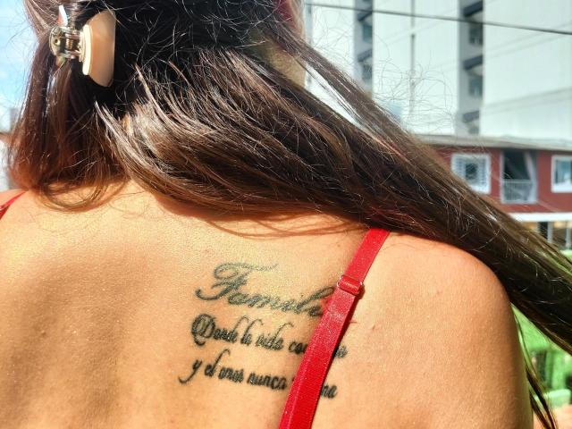 DaMikaela's bare back with a tattoo just below her red-strapped top, showing cursive text. Her brown hair is down, and she's wearing a wooden hairpin.