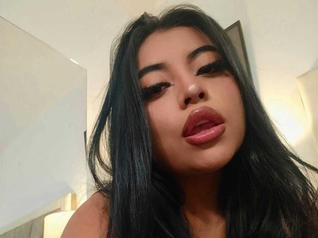 AlexaMadisson with black hair, full lips, and long eyelashes, slightly open mouth, looking down, soft lighting, visible lamp in background, warm skin tone, sexy expression.