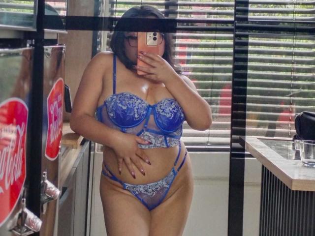 AlexaMadisson in blue lace lingerie, showing her tits and pussy in a mirror selfie, holding a phone, with one hand on her belly, in a kitchen with blinds.