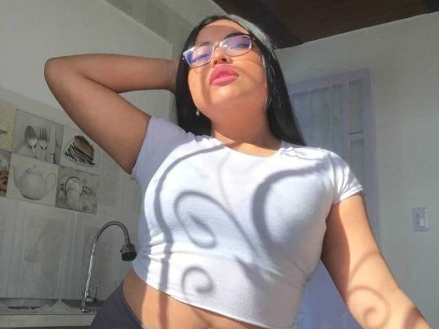 AlexaMadisson with black hair, purple glasses, and pink lipstick, wearing a white crop top showing her tits, shadow on chest, hand in hair, sunlight, kitchen background.