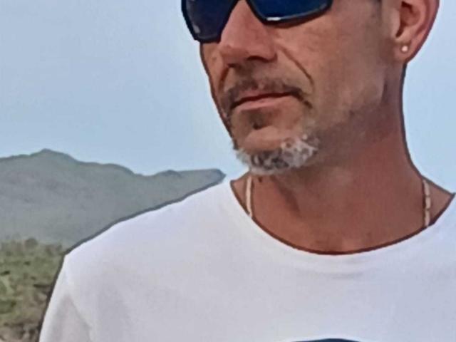 BobbyStar with a bald head, blue sunglasses, grey beard, white t-shirt, and a necklace, showing a strong jawline and a slight smile. Background is blurred.