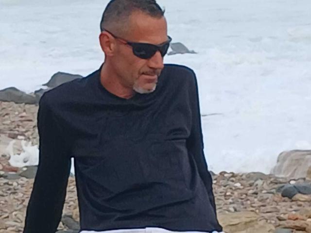 BobbyStar is a middle-aged dude with short gray hair, black sunglasses, and a black long-sleeve shirt, standing on a rocky beach, looking down with a serious face.
