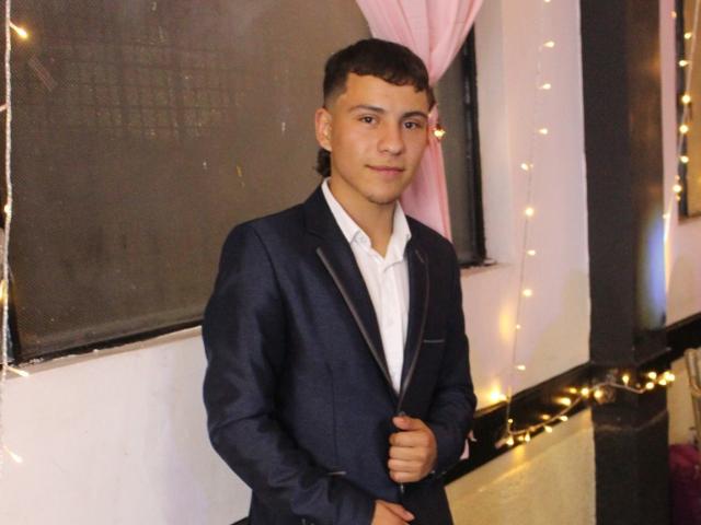 Salvador Gomez, vestito con un completo blu scuro con revers neri, camicia bianca, capelli castani corti, in piedi con le mani vicino al petto, un leggero sorriso, luci decorative sullo sfondo..