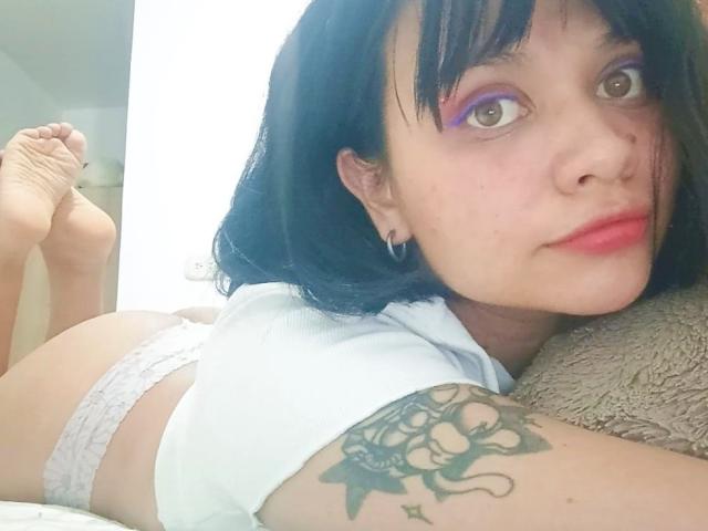 LizyOwen lying on her stomach, wearing white lace panties, showing her ass, with black hair, pink eye makeup, and a tattoo on her right arm, looking at the camera.