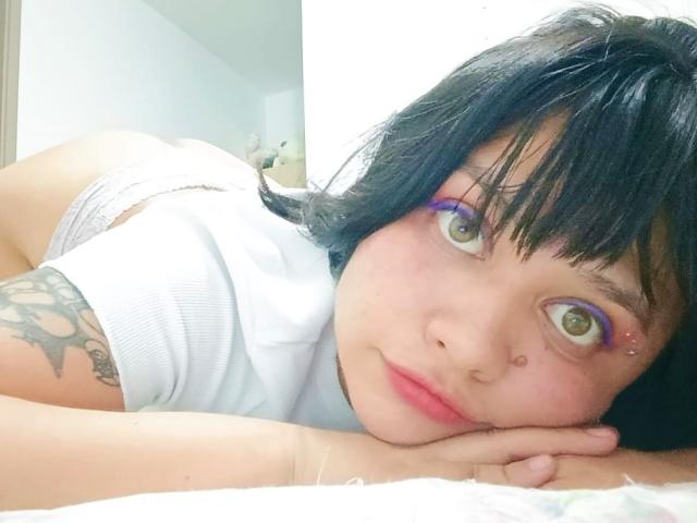 LizyOwen, with black hair, purple eyeshadow, and a nose ring, is on her bed, face down, showing her white panties and tattooed arm, with her ass raised.