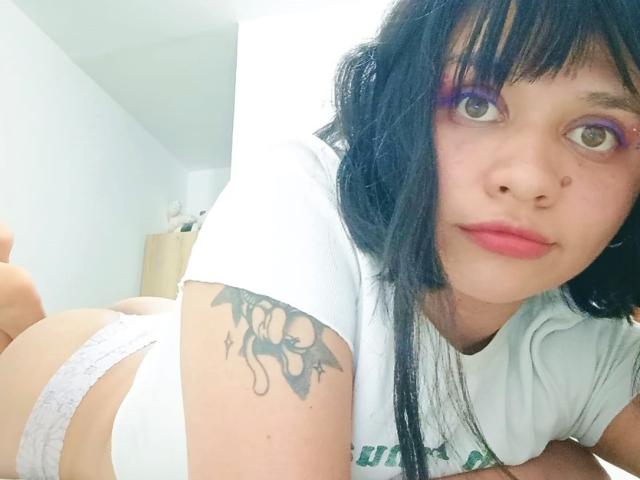 LizyOwen with black hair, purple eye makeup, white t-shirt, white lace panties, bat tattoo on arm, lying on bed, looking at camera, pink lips, pale skin, bedroom background.