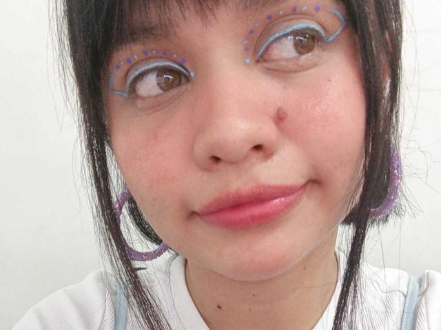 LizyOwen's face with pinkish lips, brown eyes, and blue eyeshadow with purple dots, wearing purple earrings, and a white shirt, looking up with a slight smile.