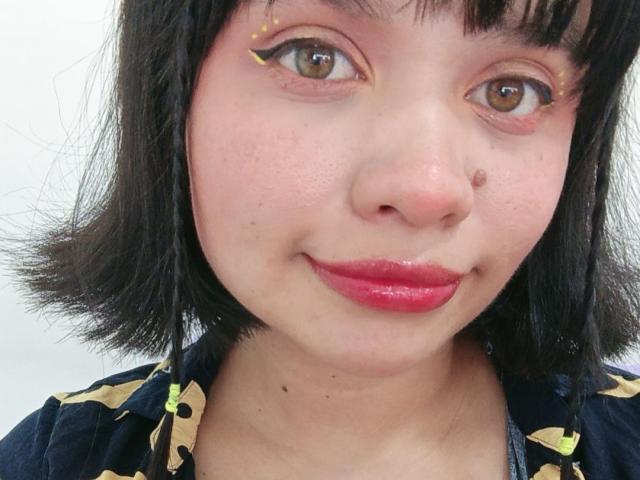 LizyOwen's face with short black hair, pinkish skin, brown eyes, yellow eyeliner, small black mole on her nose, and red lipstick. She's wearing a dark blue top with yellow patterns.