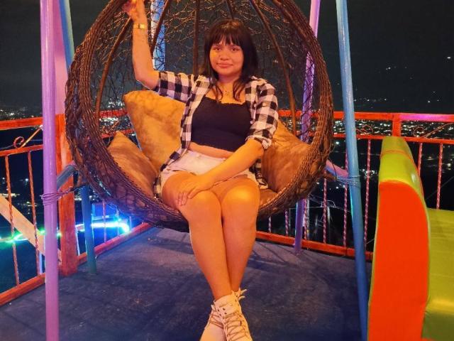 LizyOwen with medium tits, black crop top, and white shorts, sitting in a wicker swing, wearing a checkered shirt, and white sneakers, at night with city lights.