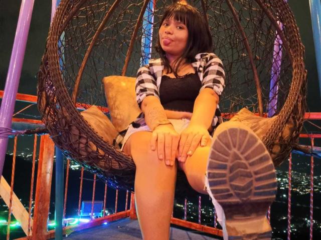 LizyOwen with black hair, puffy lips, black top, checkered shirt, white shorts, and boots, sitting in a hanging wicker chair, legs crossed, city lights in the background.