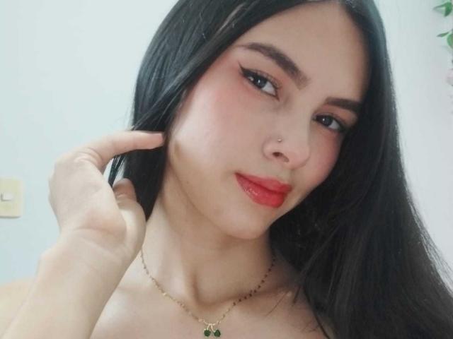 AngellAdamns has long black hair, red lipstick, and a nose piercing. She's touching her hair with her right hand, wearing a gold necklace. She's looking slightly to the side.