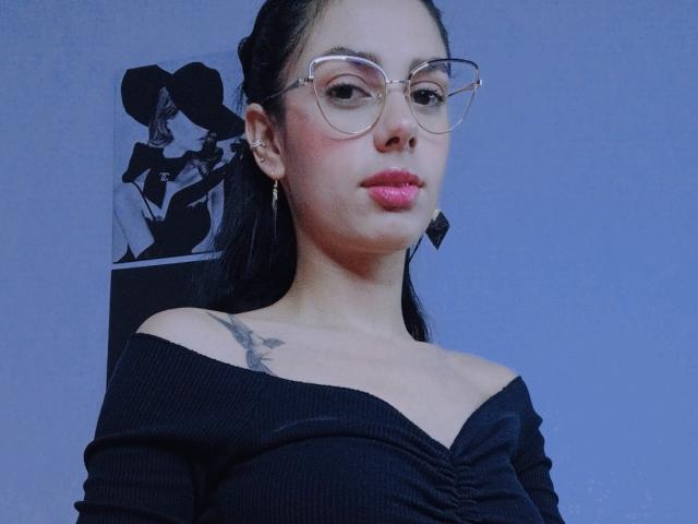 AnaisHilton with glasses, pink lips, black off-shoulder top, small tits, tattoo on left shoulder, black hair in ponytail, blue-gray background, wearing gold earrings.