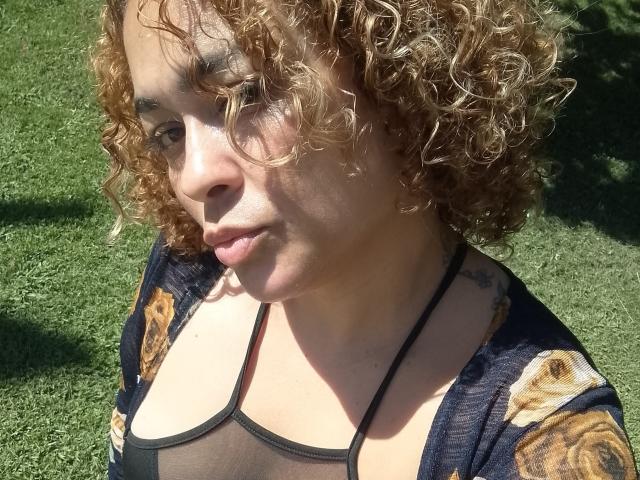 SolmaryHot with curly brown hair, light brown skin, small tattoos on shoulders, wearing a sheer black top and floral cardigan, looking slightly to the side in sunlight.