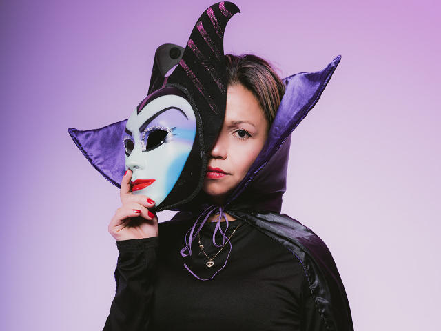 SexyKendallx wearing a black hood with purple satin trim, white mask with blue and purple accents, and red lipstick, touching mask with finger, black top, purple background.