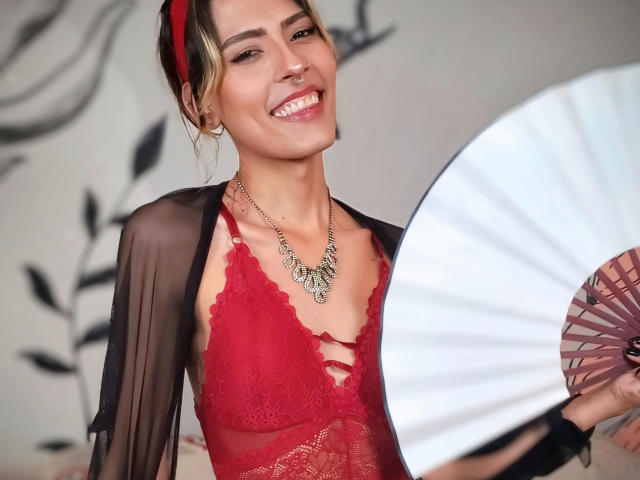 Katyling with red lace bra, sheer black robe, and necklace, holding pink fan, smiling, brown hair with red headband, small nose ring, earrings, showing off her tits.