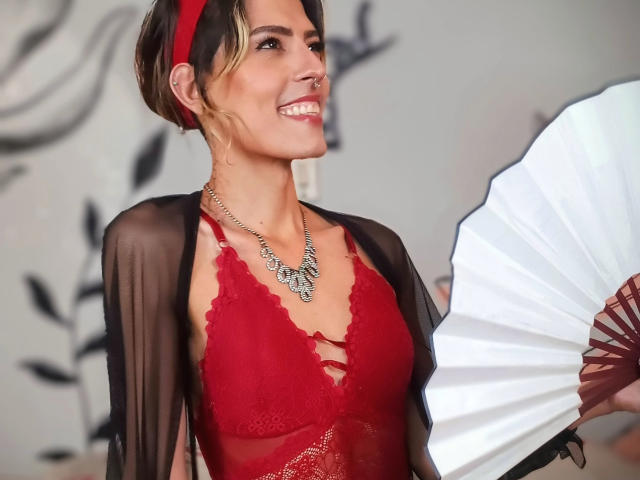 Katyling with short, blonde-streaked hair, red lace bra, sheer black robe, red headband, necklace, holding white fan, smiling, showing nose ring, small tits, attractive, confident.