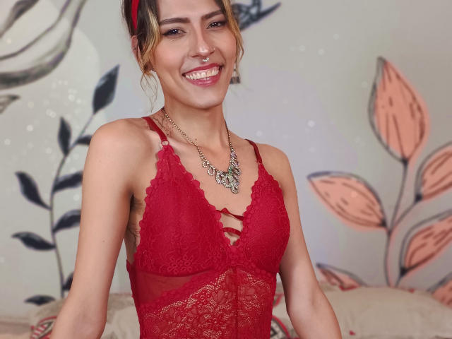 Katyling with red lace lingerie, smiling, showing her tits, has a nose ring, and a necklace with a floral design. Her hair is in a messy updo.
