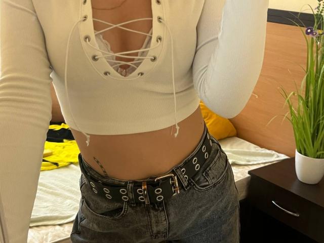 Sevillia's white, long-sleeve, lace-up top reveals her cleavage and midriff, with a black, studded belt on her blue jeans, showing a small tattoo on her side.
