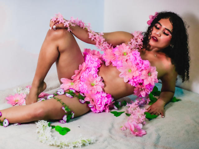 SophieSonyas with curly black hair, lying down, naked, covered in pink flowers, holding a bunch, flowers on her tits, pussy, and legs, with green leaves and white petals.