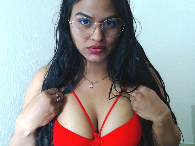 SweetAlma69 with long black hair, red bra, and glasses, holding bra straps, showing her large tits, wearing pink lipstick, and a necklace with a pink heart pendant.