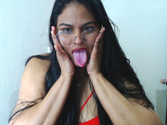 SweetAlma69 with long black hair, wearing red top, glasses, and hands on cheeks, sticking out pink tongue. Her brown eyes are focused, and she has a playful expression.