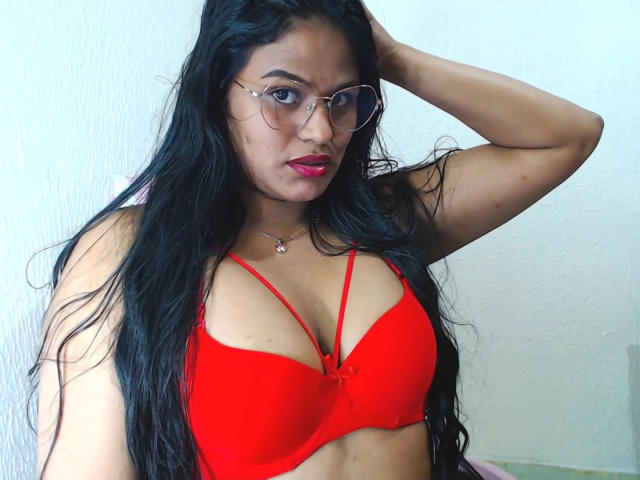 SweetAlma69 with long black hair, wearing red bra, glasses, and red lipstick, hand in hair, looking at camera, red bra highlights her tits, white wall background.