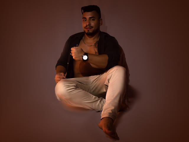 BrianMarly, with a short beard and dark hair, sits cross-legged, barefoot, wearing beige pants and an open black shirt, revealing a chest, and a watch, against a brown background.
