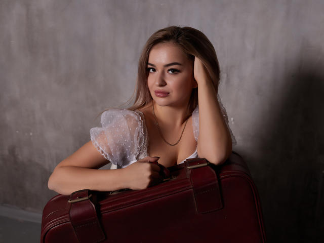 Eva Adderiy, con cabello rubio largo, vestida con una blusa blanca transparente, apoyando la cabeza en una maleta de cuero rojo, mirando a la cámara con una sonrisa sutil..