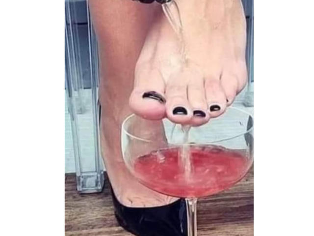 Il piede di SiPassioneX, con le dita dipinte di nero, versa una bevanda rosa in un bicchiere, la pianta del piede leggermente sopra la superficie, su un pavimento di legno..