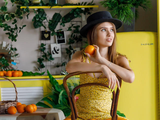 SheillaAdrian with brown hair, black hat, yellow dress, holding orange, sitting on wooden chair, yellow fridge, orange basket, green plants, looking up, medium tits, cute, sexy.