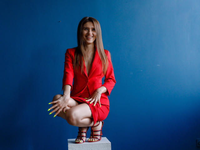 SheillaAdrian, squatting on a block, wearing a red blazer and skirt, with yellow nails, smiling, in red heels, against a blue wall, showing her tits.