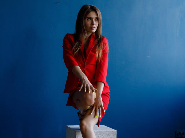 SheillaAdrian, with long brown hair, red blazer, and neon yellow nails, sits on a gray cube, legs crossed, hand on knee, against a blue wall, looking off-camera.