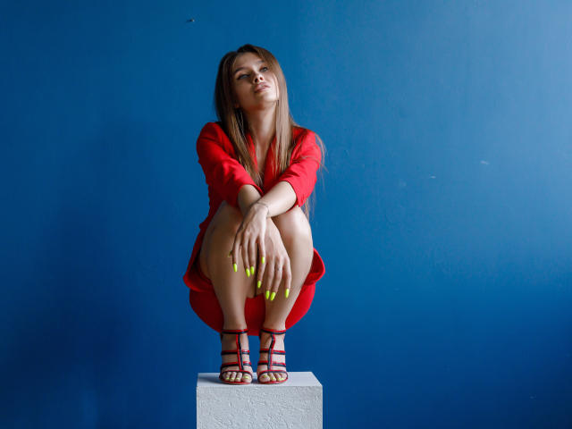SheillaAdrian, squatting on a white block, wearing a red dress, red strappy heels, and neon yellow nails, with long brown hair, and a confident, slightly smug expression.