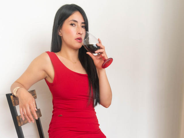 JainaRogers with long black hair, red dress, holding red wine glass, sitting on wooden chair, looking to the side, white wall background, wrist bracelet, slight pout on her face.