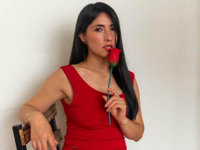 JainaRogers with long black hair, red tank top, holding red rose to her mouth, sitting on chair, looking at camera, tan skin, subtle makeup, gold bracelet, white wall.
