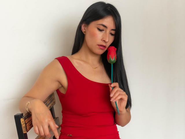 JainaRogers with long black hair, wearing a red tank top, holding a red rose to her nose, looking down, red lipstick, gold bracelet, white wall background.