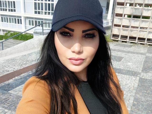 BeliseChappelleX-hot with dark eyebrows, long black hair, wearing a black cap and orange coat, has full lips, and is looking at the camera with a slight smile.
